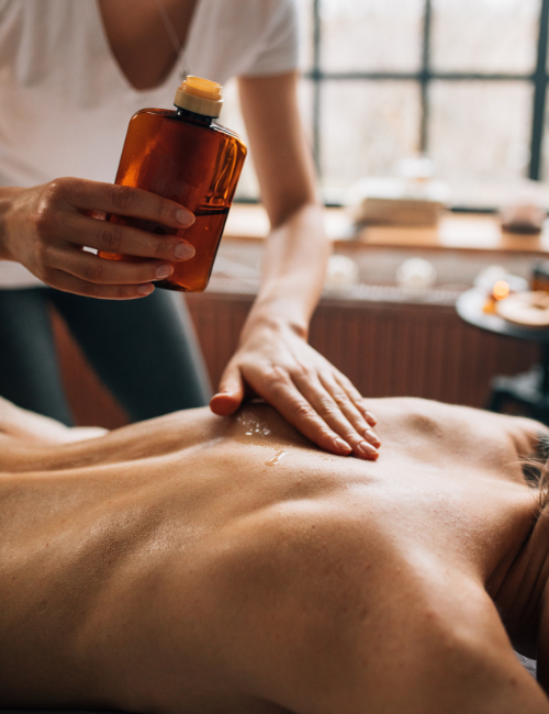 Masseur hält eine braune Flasche mit Öl in der Hand und verteilt dieses auf dem Rücken eines Kunden/in