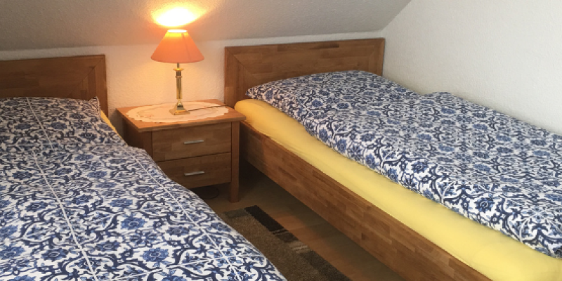 Zwei Betten in der Wohnung Klatschmohn. Gelbe Spannbetttücher und blau weiß gemusterte Bettwäsche. Eine Dachschräge am Kopfteil. Darunter ein Nachttisch mit Lampe